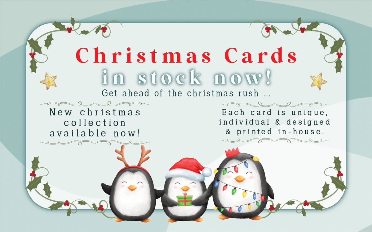 Promotional graphic for Christmas cards featuring three cartoon penguins wearing holiday accessories, with text reading 'Christmas Cards in stock now!' and decorative holly and star borders on a pale blue background.